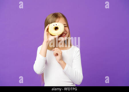 Mädchen auf lächelnde Gesicht hält süßen Donut in der Hand, violett unterlegt. Junge Mädchen mit langen Haaren gerne Süßigkeiten und Leckereien. Sweet Tooth Konzept. Mädchen auf der Suche durch die Bohrung in der Donut mit Paar Augen. Stockfoto