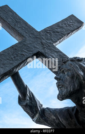 Bronzestatue von Jesus sein Kreuz trug, auf dem Weg zu seiner Kreuzigung. Ideal für Ostern, Auferstehung und andere. Blauer Himmel mit weißen cl Stockfoto