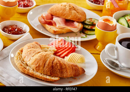 Frisch gebackene Croissant mit Butter und Erdbeere serviert mit Speck, gekochte Eier, Kaffee und Gelees oder Konfitüren für eine köstliche Frühling Frühstück Stockfoto