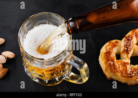 Frisches, helles Bier mit gesalzene Brezeln und Nüsse auf dem Tisch. Prozess der Gießen Sie das Getränk Stockfoto