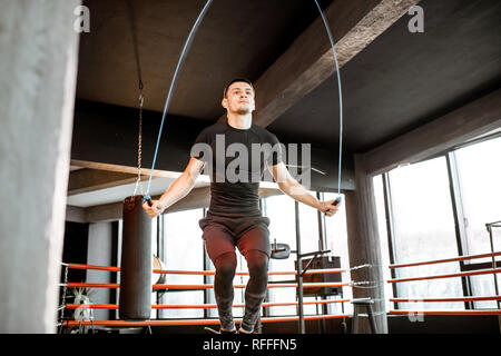 Jungen athletischen Mann in Schwarz einheitliche Ausbildung mit einem Springseil, Aufwärmen auf dem Boxring in der Turnhalle Stockfoto