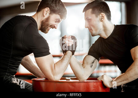 Zwei junge Athleten in schwarz Sportswear eine harte Arm Wrestling Wettbewerb auf einem roten Zylinder in der Turnhalle Stockfoto