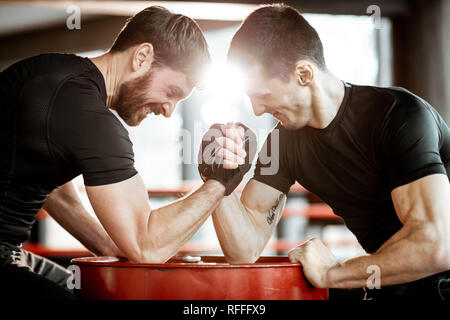 Zwei junge Athleten in schwarz Sportswear eine harte Arm Wrestling Wettbewerb auf einem roten Zylinder in der Turnhalle Stockfoto