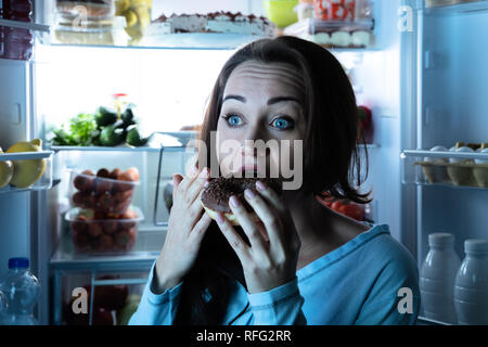 Nahaufnahme einer jungen Frau Essen Donut Vor dem offenen Kühlschrank Stockfoto