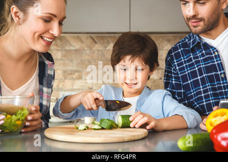 Familie zu Hause in der Küche zusammen Mutter und Vater auf der Suche nach dem Sohn schneiden Gurke in der Nähe konzentrierten Stockfoto