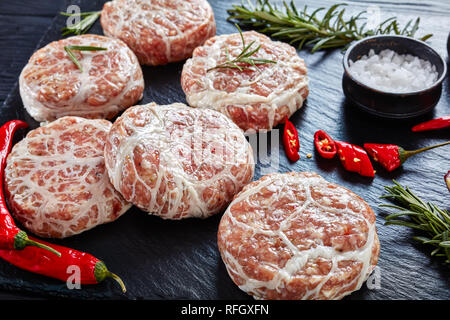 Raw Atriaux - Schweinefleisch in caul Fett gewickelt, Schweizer Küche, Art der Bratwurst, Hamburger auf einer Schiefertafel Fach mit Zwiebel, Rosmarin, Pfeffer und Salz, o Stockfoto