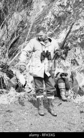 Schwarz-weiß Foto von einem Mann mittleren Alters stehen in voller Länge ansehen und trug eine Jagd Jacke, hohe Schnürschuhe Stiefel und Handschuhe, mit dem Fernglas mit Schlingen um den Hals und ein Gewehr in der Hand, auf der Suche nach einem toten Vogel in der anderen Hand gehalten, mit zwei weiteren Zahlen, die hinter ihm saßen auf dem Boden und schaut durch ein Fernglas, mit einem Felsen oder steilen Hang im Hintergrund sichtbar, während einer Jagd und Angeln in Alaska, USA, 1955 entfernt fotografiert. () Stockfoto