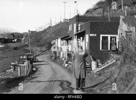 Schwarz-weiß Foto eines Mannes, von der Rückseite aus betrachtet, trägt einen langen Mantel und stehen am Rand der Piste, mit seinen Armen, hob den Kopf (wahrscheinlich mit einer Kamera) Mit einer Küstenlinie und alten Auto auf der linken Seite, mehrere kleine Gebäude aus Holz (mit Zimmer die Zeichen '' und 'Yum Yum isst") an der rechten und mehr Gebäude aus Holz und die schneebedeckten Berge im Hintergrund sichtbar, während einer Jagd und Angeln in Alaska, USA, 1955 entfernt fotografiert. () Stockfoto