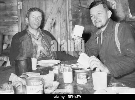 Schwarz-weiß Foto von zwei unrasiert, Männer im mittleren Alter, jedem tragen wollene Shirts und Hosenträgern, sitzen gemeinsam in einer Blockhütte oder Shack, essen eine Mahlzeit in einem unordentlichen Tabelle, mit verschüttetem Zutaten, ein Glas Milch oder Zucker, und eine Dose "Marke Pfirsiche York' im Vordergrund sichtbar, während einer Jagd und Angeln in Alaska, USA, 1955 entfernt fotografiert. () Stockfoto