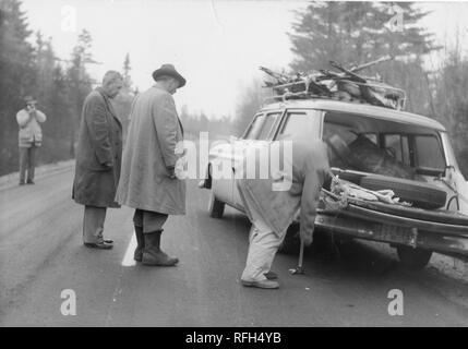 Schwarz-weiß Foto von vier Männern, jeder trägt einen Mantel oder Jacke, die auf einem leeren Strecke von der Straße mit einem geparkten Kombi, die über die Körper von mehreren Hirsch in seiner overhead Rack; ein Mann im Vordergrund rechts zu sein scheint, ändern der hinteren Reifen während zwei weitere Männer ihn beobachten, der vierte Mann Filme oder Fotos im Verfahren von der linken Hintergrund; mit Koniferen und andere Bäume im Hintergrund sichtbar, während einer Jagd und Angeln in Alaska, USA, 1955 entfernt fotografiert. () Stockfoto