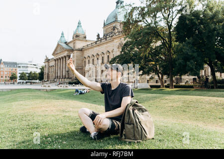 Eine junge männliche Touristen oder Studenten sitzt auf dem Gras außerhalb in Leipzig, Deutschland, sprechen auf Video oder mit dem Handy zu chatten, Bilder oder. Stockfoto