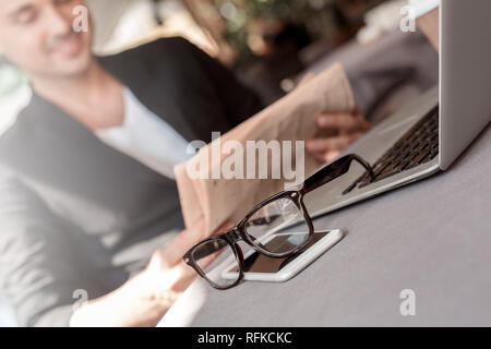 Mittagessen Zeit. Junger Mann in Anzug im Cafe am Ort Straße lesen Zeitung freudigen Sitzung verschwommen Brillen und Telefon close-up Stockfoto