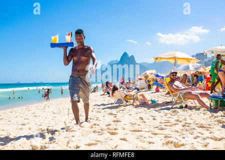 RIO DE JANEIRO - Februar, 2018: Eine nicht lizenzierte brasilianischen Strand Verkäufer trägt ein Tablett mit hausgemachten Caipirinha cocktails Vergangenheit Badegäste am Strand von Ipanema Stockfoto