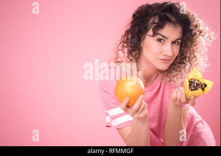 Portrait einer jungen lockigen dunkelhaarigen Mädchen bei der Wahl der richtigen Nahrungsmittel mit Orange und Kuchen in der Hand, in einem losen rosa T-Shirt isoliert auf einem Rosa backgro Stockfoto