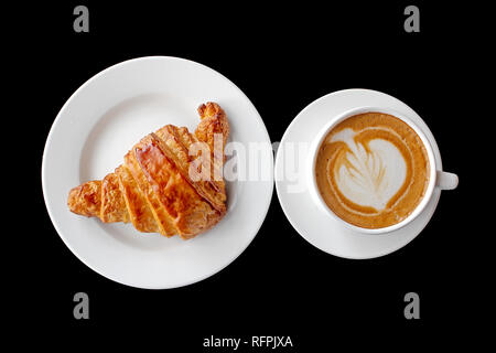 Ein Cappuccino und ein Croissant isoliert auf schwarzem Hintergrund. Leichtes Frühstück, Blick von oben. Stockfoto