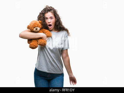 Junge brünette Mädchen, dass Teddybären über isolierte Hintergrund im Schlag mit einer Überraschung konfrontiert, ängstlich und aufgeregt mit Angst Angst Ausdruck Stockfoto