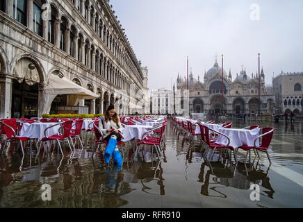 Eine Frau sitzt auf einem Stuhl, der auf Restaurant San Marco Platz, Piazza San Marco, während der Acqua Alta überflutet Stockfoto