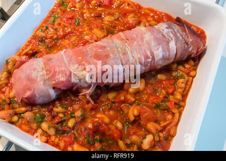 Close up Italienische Gericht aus Schweinefilet in Prosciutto eingewickelt bereit zum Kochen auf Bett von Tomaten, Bohnen, Kräutern und Knoblauch in weißen Keramikschale Stockfoto