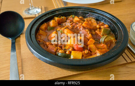 Vegetarisches Essen von Gemüse Tajine aus Gusseisen Topf mit Kürbis, Zucchini, Zwiebeln, Paprika und Kelle auf Eiche Tisch Stockfoto