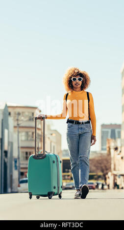 Afro-amerikanische Frau zu Fuß auf der Straße mit einem Gepäckwagen. Frau in Sonnenbrille trägt einen Rucksack und Trolley Tasche zu Fuß rund um den Stockfoto