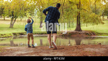 Ansicht der Rückseite des Vater und Sohn werfen Steine in den Teich im Park. Mann und kleinen Jungen Spaß im Park. Kind mit Vater werfen Steine in den Teich. Stockfoto