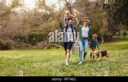 Glückliches Paar mit ihren zwei Kindern und Hund Wandern im Park. Mann, der kleine Mädchen mit Drachen auf Schultern und Frau, die einen Picknickkorb mit Stockfoto