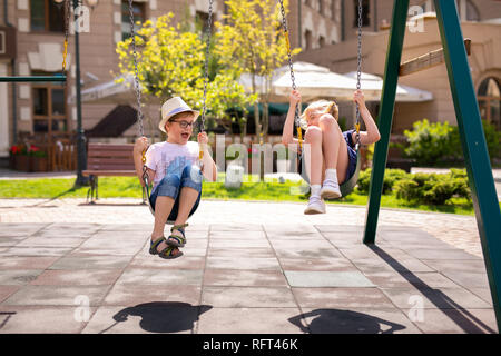 Junge in die Brille und Hut und Blonde Mädchen im Kleid Spaß auf einer Schaukel zusammen in schönen Sommergarten an warmen und sonnigen Tag im Freien. Stockfoto