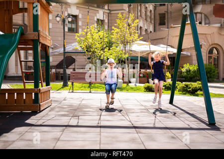 Junge in die Brille und Hut und Blonde Mädchen im Kleid Spaß auf einer Schaukel zusammen in schönen Sommergarten an warmen und sonnigen Tag im Freien. Stockfoto