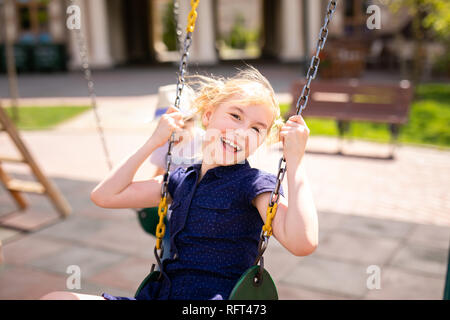 Junge in die Brille und Hut und Blonde Mädchen im Kleid Spaß auf einer Schaukel zusammen in schönen Sommergarten an warmen und sonnigen Tag im Freien. Stockfoto