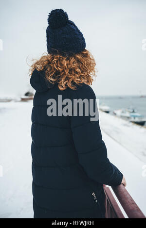 Rückansicht der jungen Frau mit warmen Mantel und Strickmütze stehend auf verschneiten Deich alleine am Meer im Winter. Ehrliches Bild der Rothaarige Stockfoto