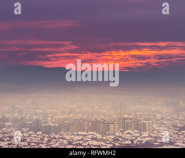 Erstaunlich brennenden Sonnenuntergang Himmel über Diesig, neblig, goldene Stadtbild von Pirot mit rosa und lila Himmel und Gebäude im Smog bedeckt Stockfoto