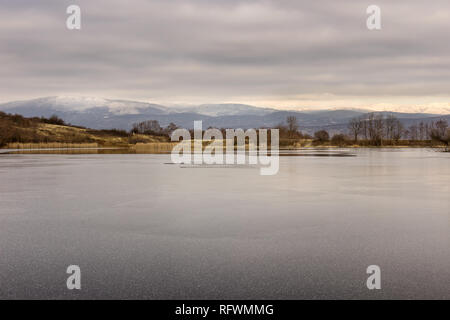 Blick über wunderschöne, halb zugefrorenen See mit Schneeflocken auf neue Eis- und Reed und die schneebedeckten Berge von der anderen Seite Stockfoto