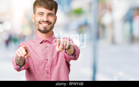 Jungen gutaussehenden Mann mit rosa Shirt über isolierte Hintergrund zeigt auf sie und die Kamera mit den Fingern, Lächeln, positiven und fröhlichen Stockfoto