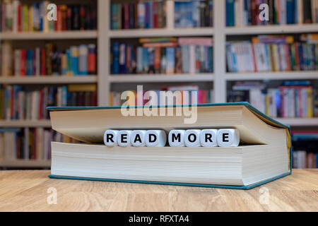 Würfel in Buchform, die Wörter "Anzeige mehr'. Buch liegt auf einem hölzernen Schreibtisch vor einem Bücherregal. Stockfoto