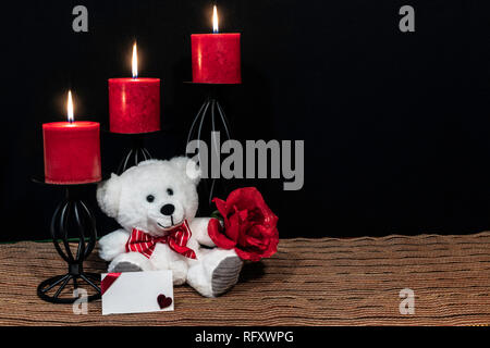 Cudlely Teddybär mit roter Schleife binden, rote Rose, rote Kerzen thront auf schwarz Kerzenhalter auf Netz Unterlage und Holztisch mit Karte und dunklen zurück Stockfoto
