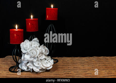 Schöne bouquie der weißen Rosen, rote Kerzen thront auf schwarz Kerzenhalter auf Netz Unterlage und Holztisch mit Karte und dunklen Hintergrund. Valent Stockfoto