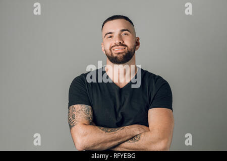 Junge starke glücklich bärtiger Mann mit verschränkten Armen auf grauem Hintergrund Stockfoto