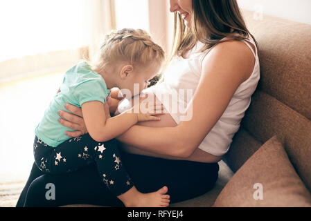 Seitenansicht der glücklichen schwangeren Mutter sitzt auf einem Sofa und niedlichen Tochter küssen und umarmen großen Bauch. Schöne Mädchen erwarten kleiner Bruder. Konzept der Stockfoto