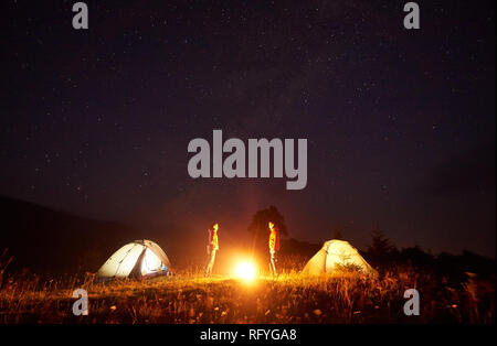 Nacht Camping. Hellen Lagerfeuer brennen zwischen zwei Touristen, Junge und Mädchen stehen einander gegenüber Vor beleuchtete Zelte unter schönen da Stockfoto