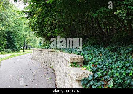 Die Straße geht in die Ferne und dreht sich hinter einem Stein Zaun. Dick grün Efeu Winde im Schatten der Bäume über dem Zaun Stockfoto