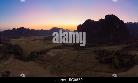 Antenne: Vang Vieng backpacker Reiseziel in Laos, Asien. Sonnenuntergang über der malerischen Klippen und Felsen Pinnacles, Reisfeldern Tal, atemberaubende Landschaft. Stockfoto
