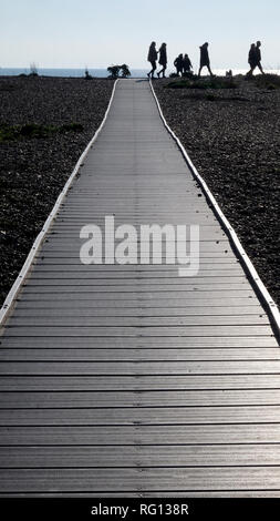 Sieben silhouetted unkenntlich Menschen zu Fuß über das Ende eines langen Geraden Holz- weg mit einem Kieselstrand auf beiden Seiten eine kleine Linie des Meeres Stockfoto