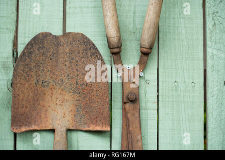 Alte verrostete gardening Tools, einschließlich einer Schaufel und Gartenscheren auf einem Holz Garten Zaun montiert grün lackiert Stockfoto