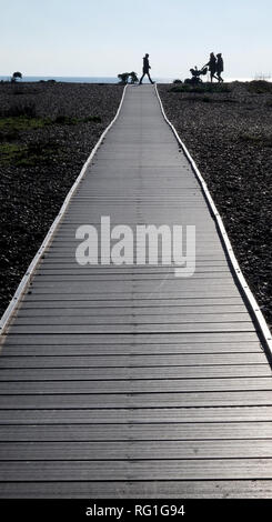 Drei silhouetted unerkennbare Personen und ein Baby in einem Kinderwagen zu Fuß über das Ende eines langen Geraden Holz- weg mit einem Kieselstrand auf beiden Seiten Stockfoto