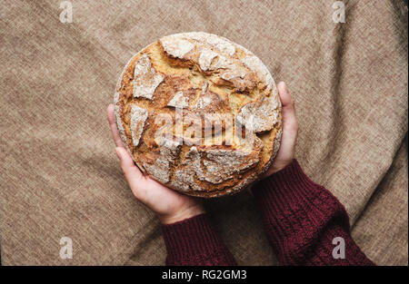 Frau Hand, braunes Brot von Oben nach Unten anzeigen. Gebackene ganzen Roggenkorn hausgemachte Runde Laib auf rustikalen Leinen Sack Hintergrund. Baker Lebensmittel frisch Runde H Stockfoto