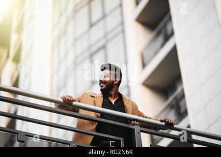 Low Angle View Porträt einer indischen Mann mit Sonnenbrille und Jacke beim Tagträumen im Freien in einer modernen Stadt Stockfoto