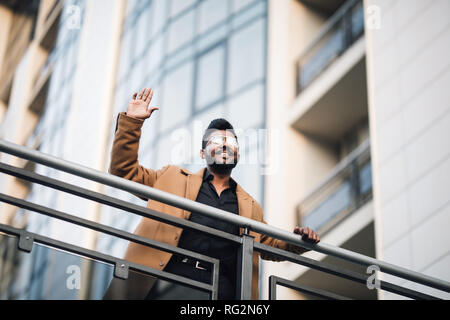 Ernsthafte indischer Geschäftsmann in Brillen suchen auf Kopieren, während sich gegen Glashochhaus winken Hallo zu empfehlen. Stockfoto