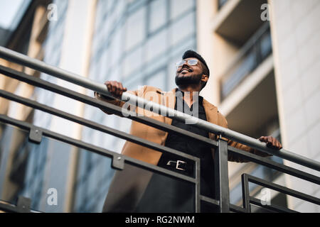 Low Angle View Porträt einer indischen Mann mit Sonnenbrille und Jacke beim Tagträumen im Freien in einer modernen Stadt Stockfoto