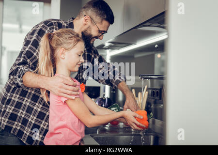 Langhaarige hübsches Mädchen in einem rosa Hemd und ihr Vater Waschen von Gemüse in der Küche Stockfoto