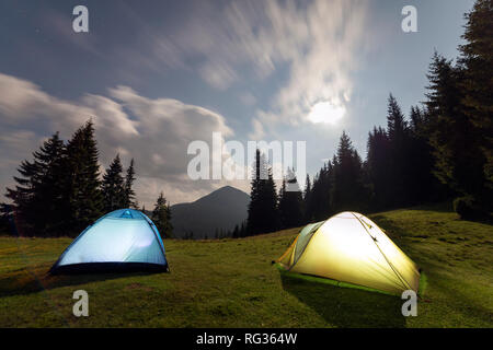 Hellen Mond in Dunkelblau bewölkter Himmel über zwei touristische Zelte auf der grünen Wiese Wald Lichtung unter hohen Kiefern auf ferne Berge im Hintergrund. T Stockfoto